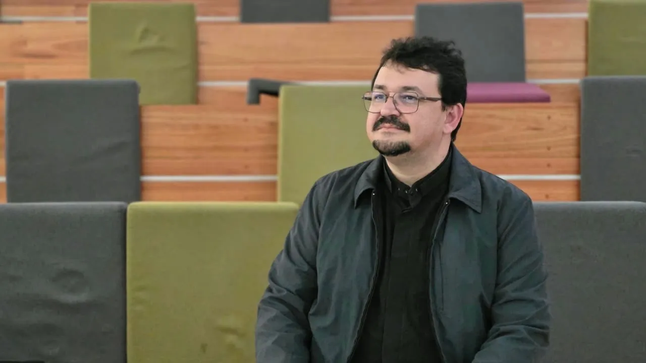 Padre Jocleilson paramentado de roxo, de mãos unidas, diante de uma grande janela clara
